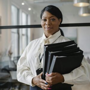 Smiling African American female employee wearing classy outfit standing with paper documents in modern workplace